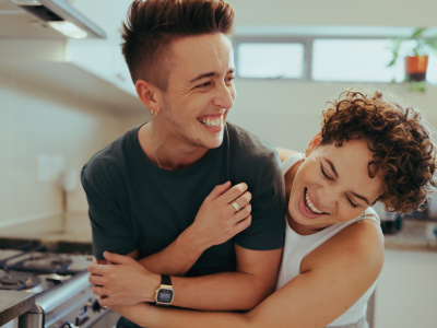 Young queer couple laughing together indoors. Happy young queer couple having fun together while standing in their kitchen. Romantic young LGBTQ+ couple bonding fondly at home.