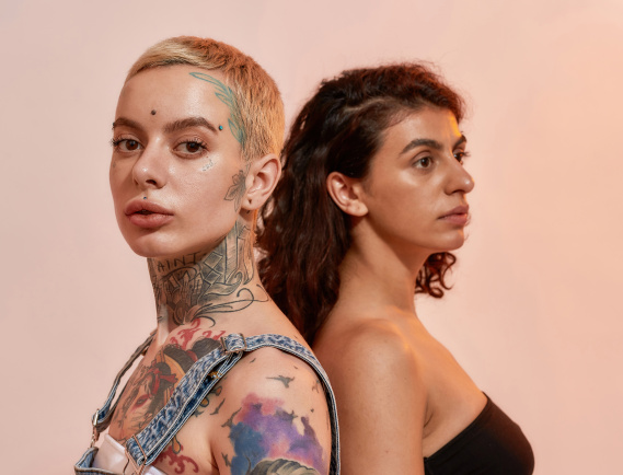 Two young diverse lesbian women standing back to back, posing together isolated over light pink background. Lgbtq, youth concept. Web Banner