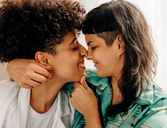 Playful lesbian couple touching their noses together at home. Two affectionate female lovers flirting with each other. Romantic young lesbian couple sitting together in their living room.